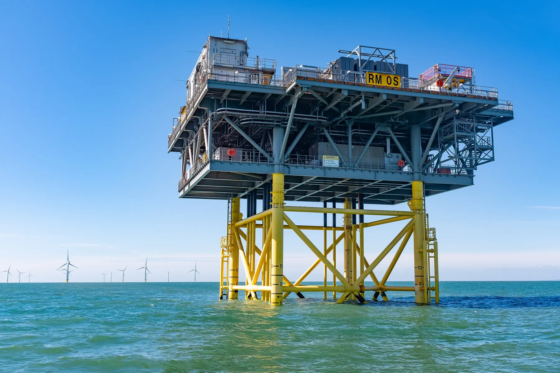 An offshore wind farm substation platform standing on yellow legs in open sea, turbines behind