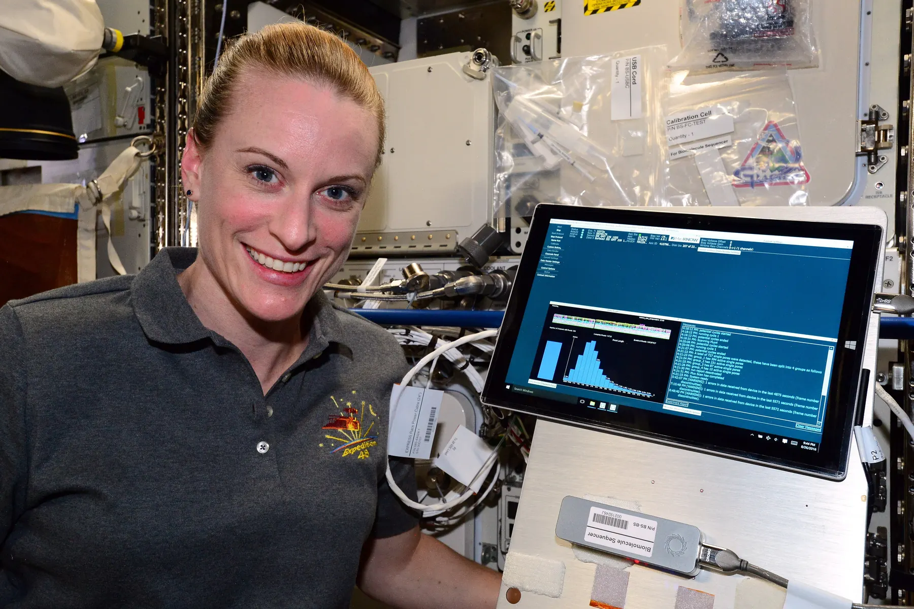 An astronaut aboard the space station holding a small handheld DNA sequencer beside a laptop